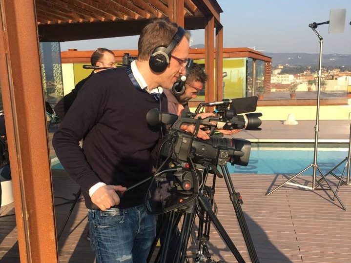 Un operatore video con cuffie sta preparando una telecamera su un terrazzo soleggiato, mentre altri membri del team lavorano in background.