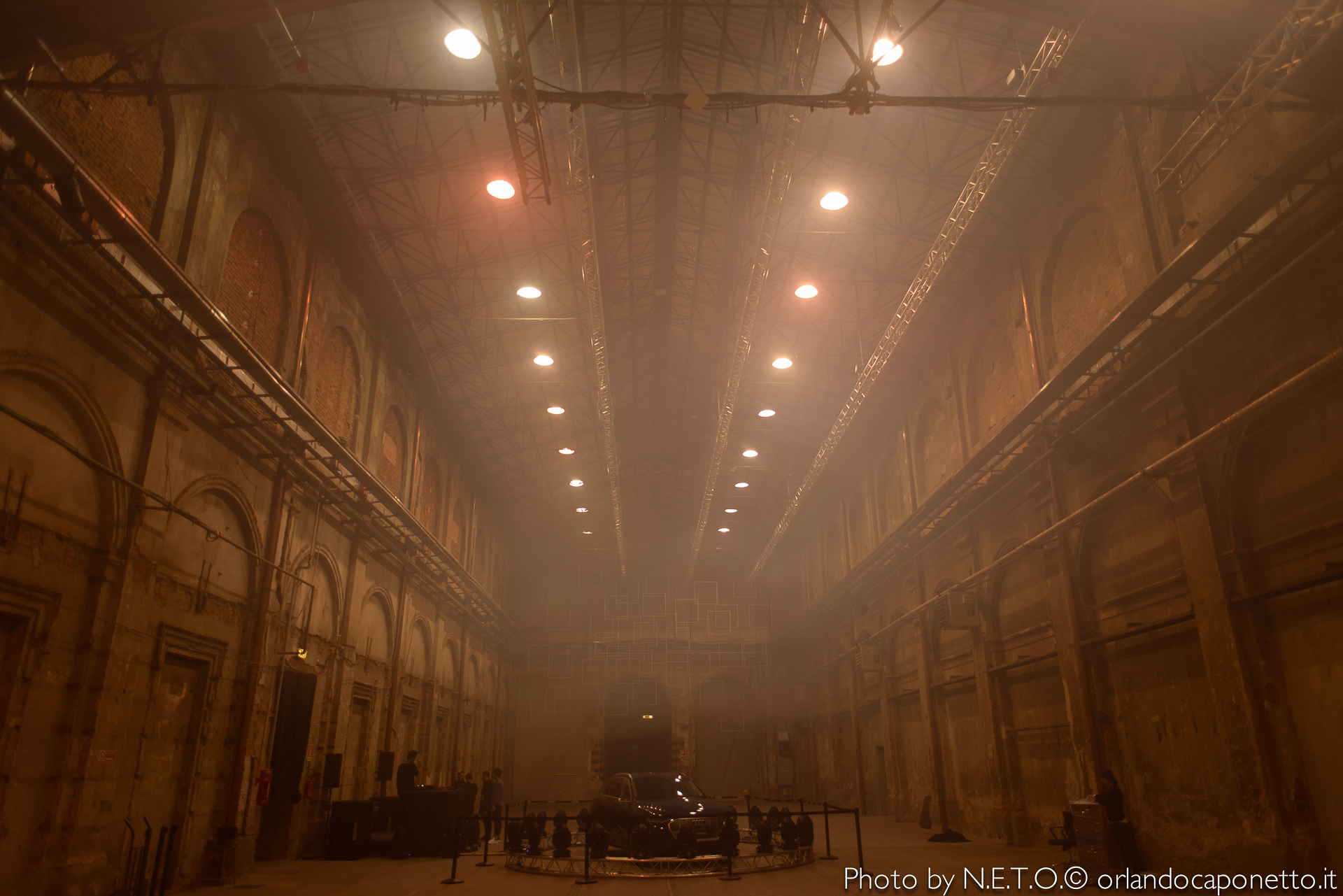 Interno di un grande spazio industriale con nebbia leggera e illuminazione sospesa, un'auto nera al centro circondata da attrezzature e persone.