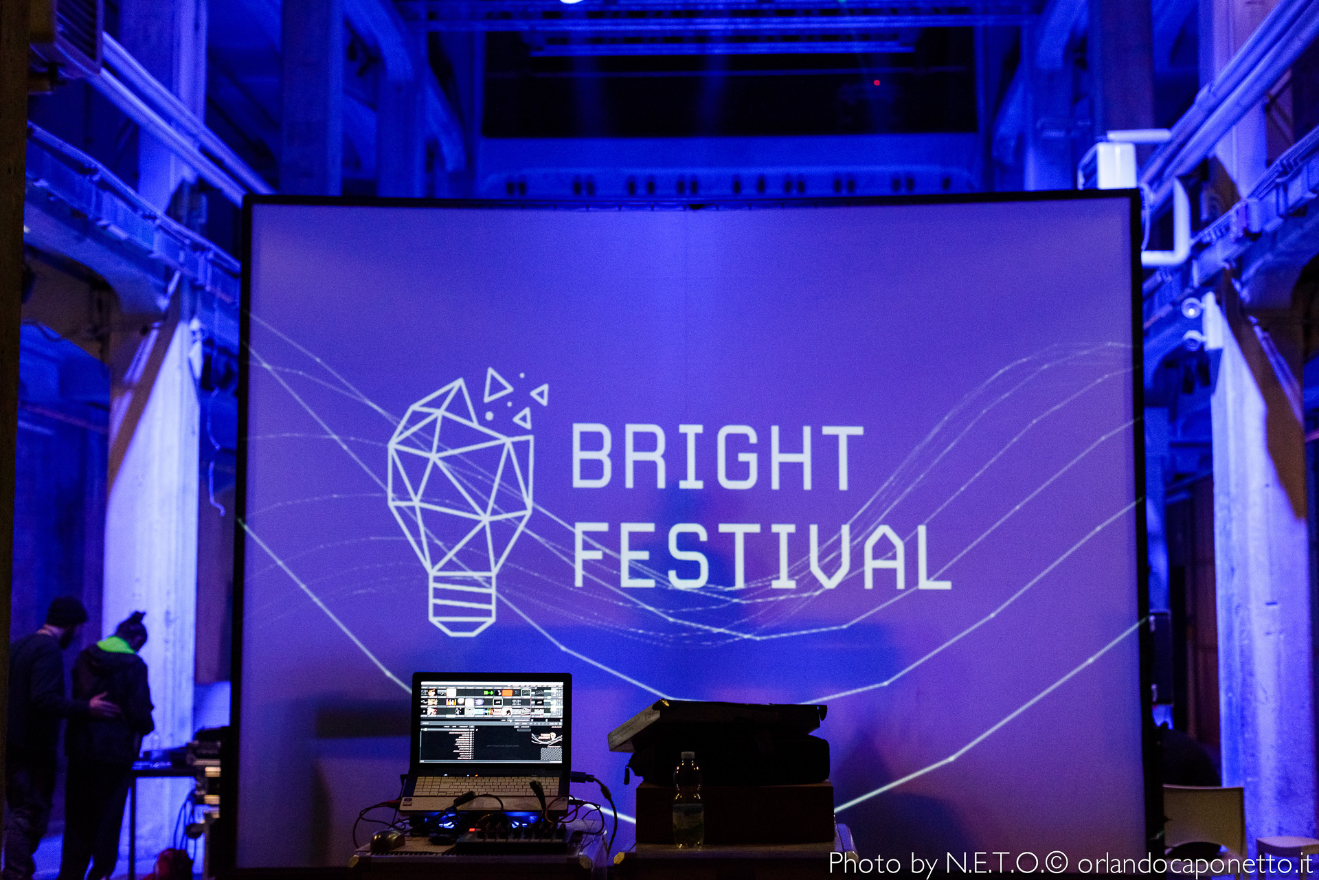 Schermo che presenta il logo del Bright Festival, con un computer portatile in primo piano, situato in un ambiente illuminato da luci blu.