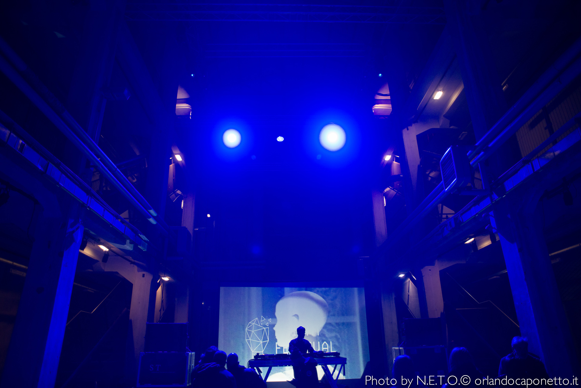 Un DJ si esibisce sotto luci blu in un ambiente di concerto, con uno schermo sullo sfondo che mostra un'immagine stilizzata di un volto umano.