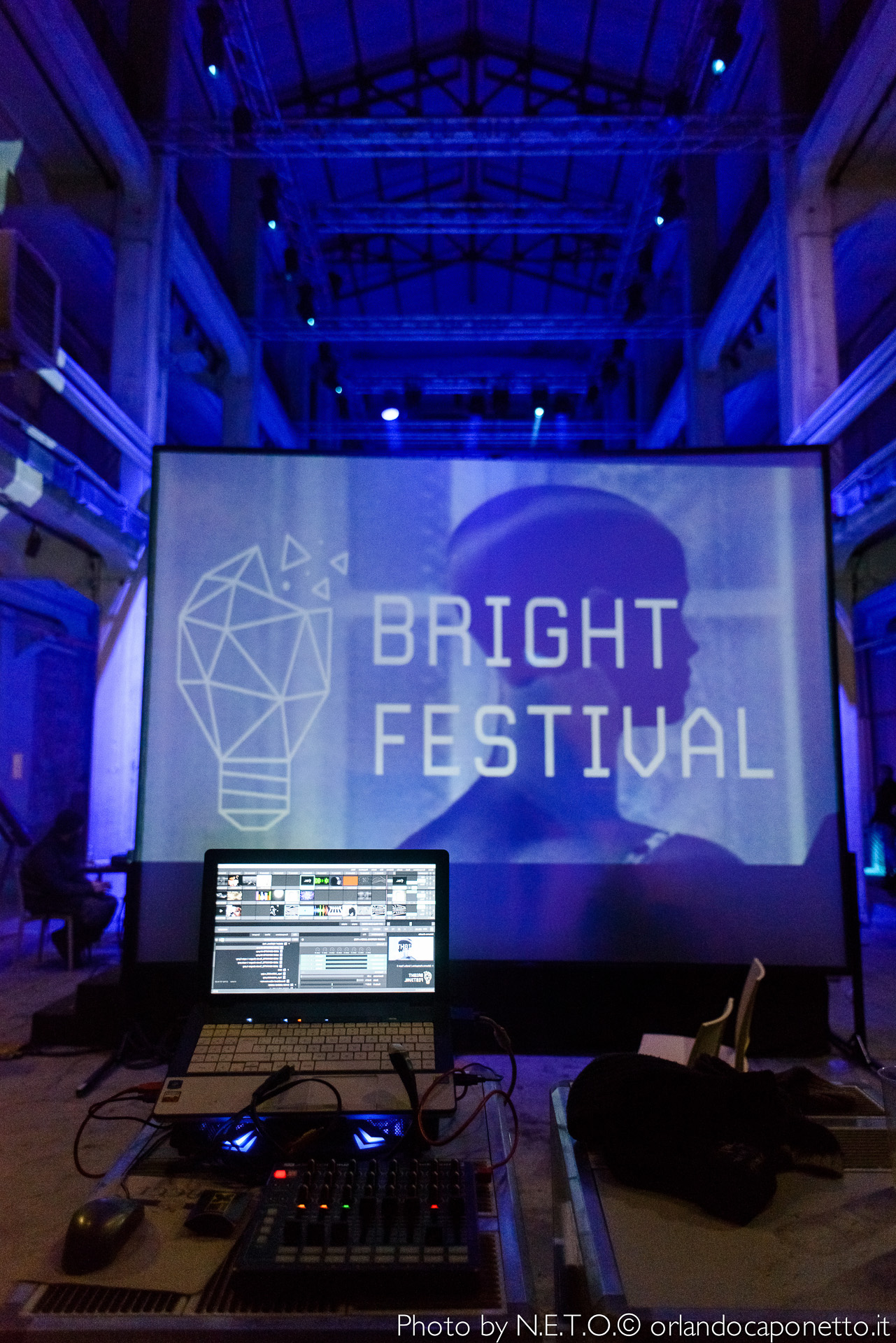 Vista di un evento del Bright Festival, con un proiettore che mostra il logo del festival su uno schermo grande sullo sfondo e un computer con attrezzature audio in primo piano.