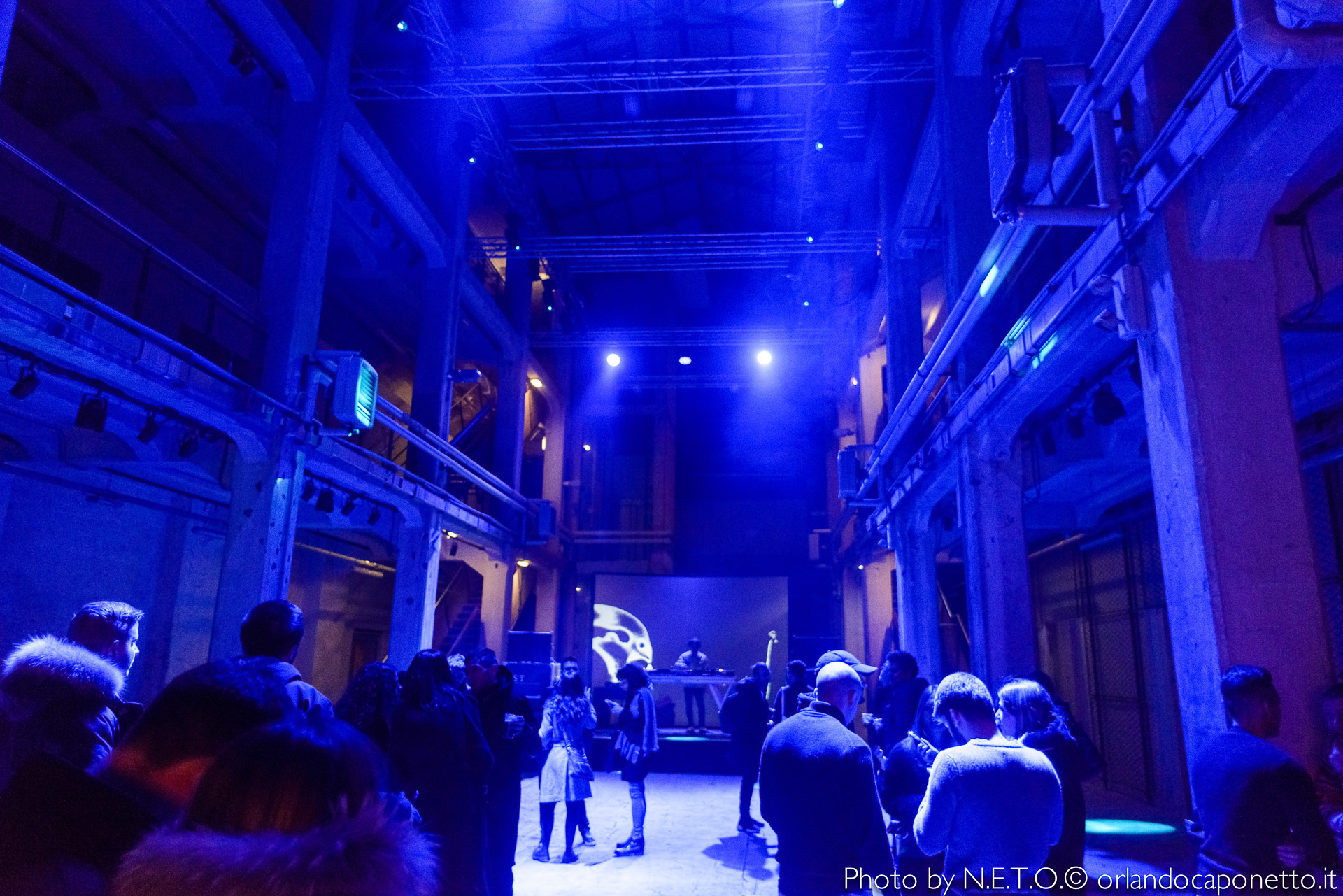 Interno di un locale con luci blu soffuse e fumi, dove si vedono persone che interagiscono, davanti a un DJ e a uno schermo proiettato.
