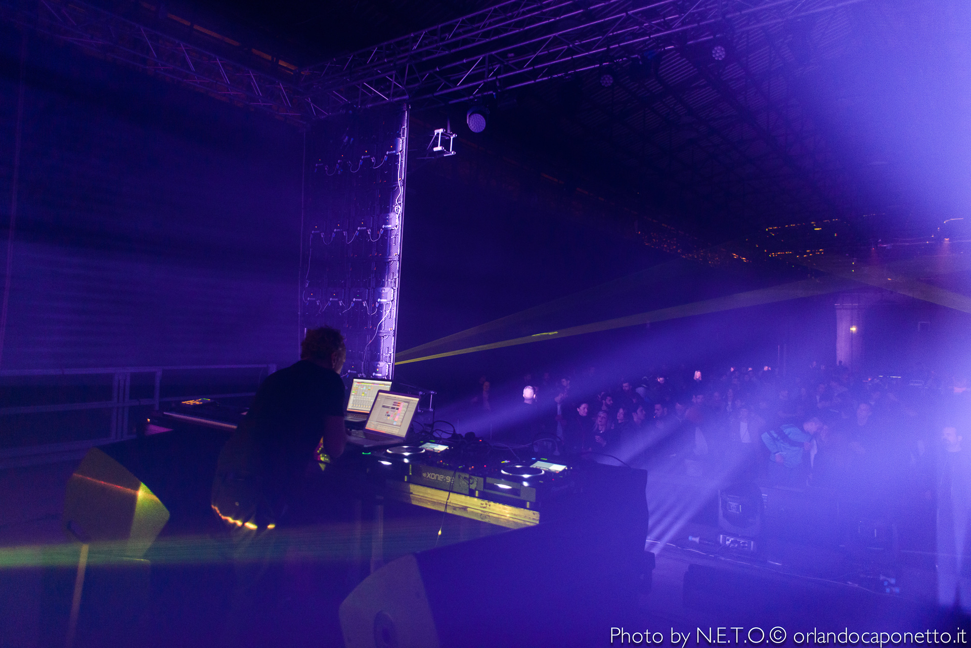 Un DJ che suona alla console con luci viola e raggi laser, davanti a una folla di persone in un ambiente di festa.