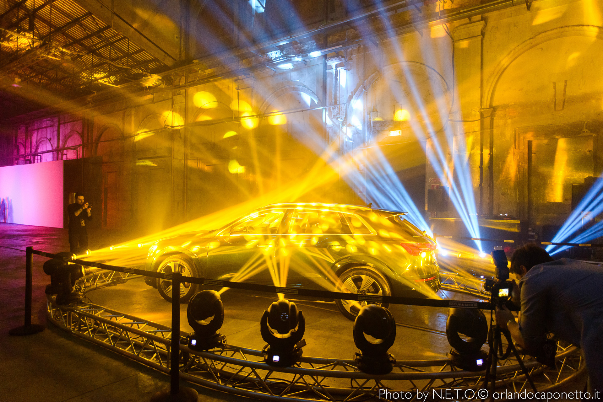 Un'auto esposta in un evento con luci dorate e blu che illuminano l'ambiente, mentre un fotografo scatta foto da un lato.