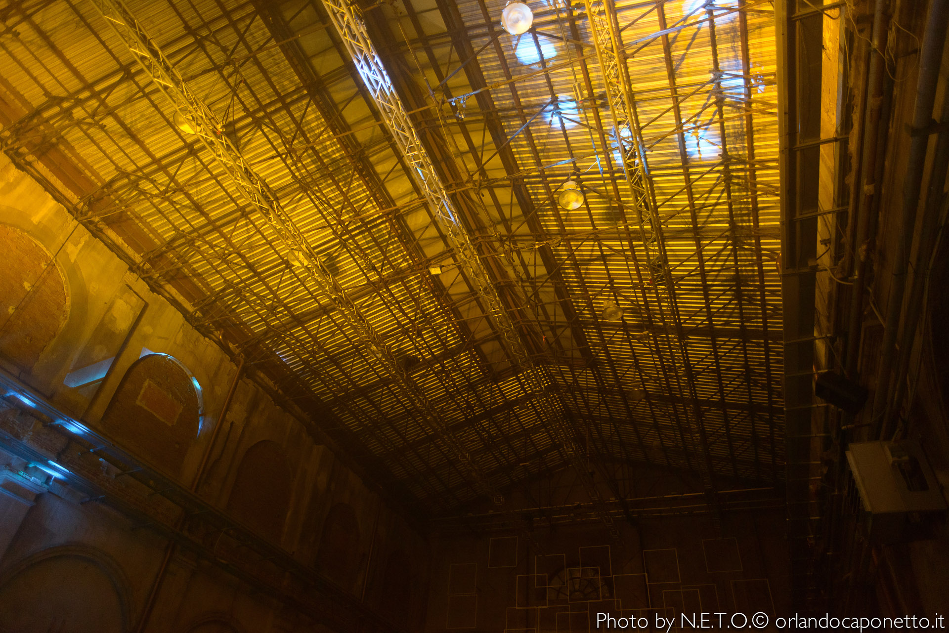 Soffitto di un grande edificio industriale con strutture metalliche e luci gialle, visibile dall'alto.