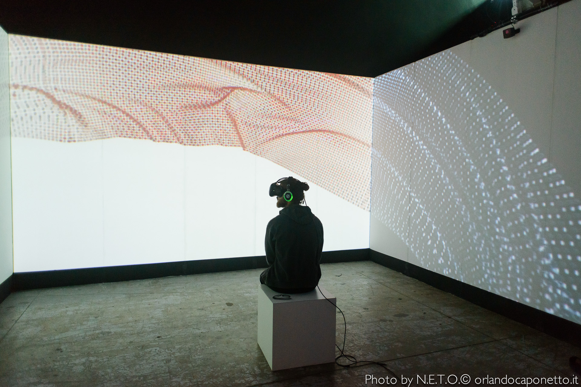 Una persona seduta su un cubo bianco indossa un visore VR in una stanza. Le pareti sono decorate con proiezioni di colori e forme astratte.