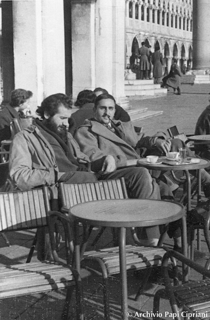 Due uomini (Emilio Vedova e Giorgio Cipriani) seduti a un tavolo all'aperto, in una piazza, con una tazza di caffè e un'atmosfera rilassata.