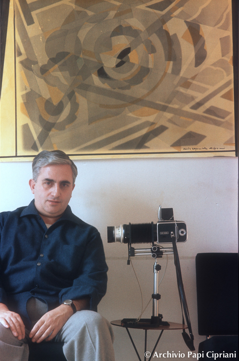 Un uomo, Giorgio Cipriani, seduto accanto a una macchina fotografica vintage, con un dipinto astratto sulla parete sullo sfondo.