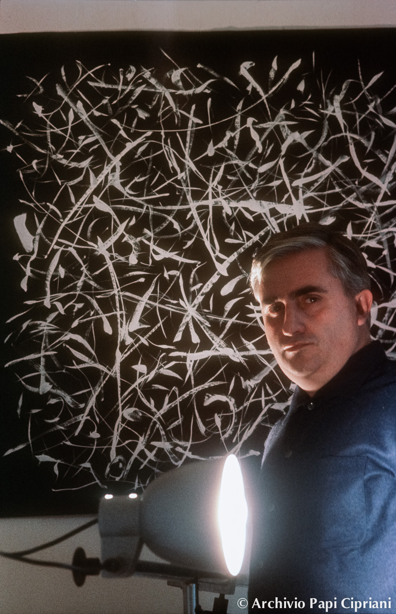 Un uomo in primo piano di fronte a un'opera d'arte astratta su sfondo scuro, con linee bianche intrecciate. Una lampada illumina il suo volto.