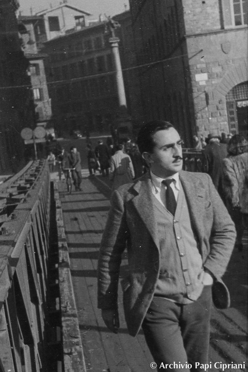 Un uomo vestito elegantemente cammina su un ponte, circondato da altre persone e edifici storici in una città italiana.
